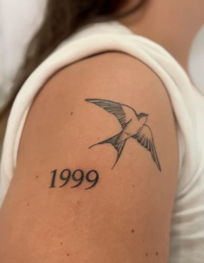 Tatouage oiseau avec la date 1999 tatoué sur le haut du bras d'une femme - Tattoo chez L' (Vonnas)